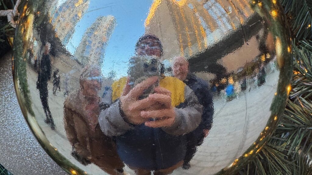 Distorted fisheye reflection in a gold Christmas tree bauble showing a person holding a phone taking a selfie, with Battersea Power Station's iconic chimneys and blue sky visible in the curved reflection, framed by green pine needles