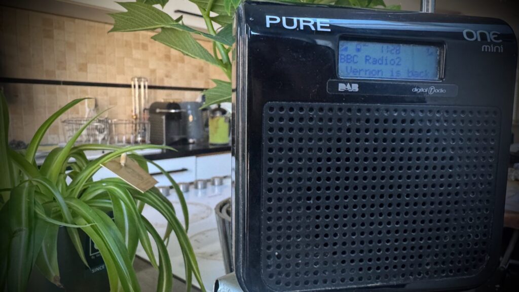 Black Pure One Mini DAB digital radio displaying 'BBC Radio2' and 'Vernon is here' on its screen, positioned next to a spider plant in a modern kitchen with cream tiled splashback and white worktop