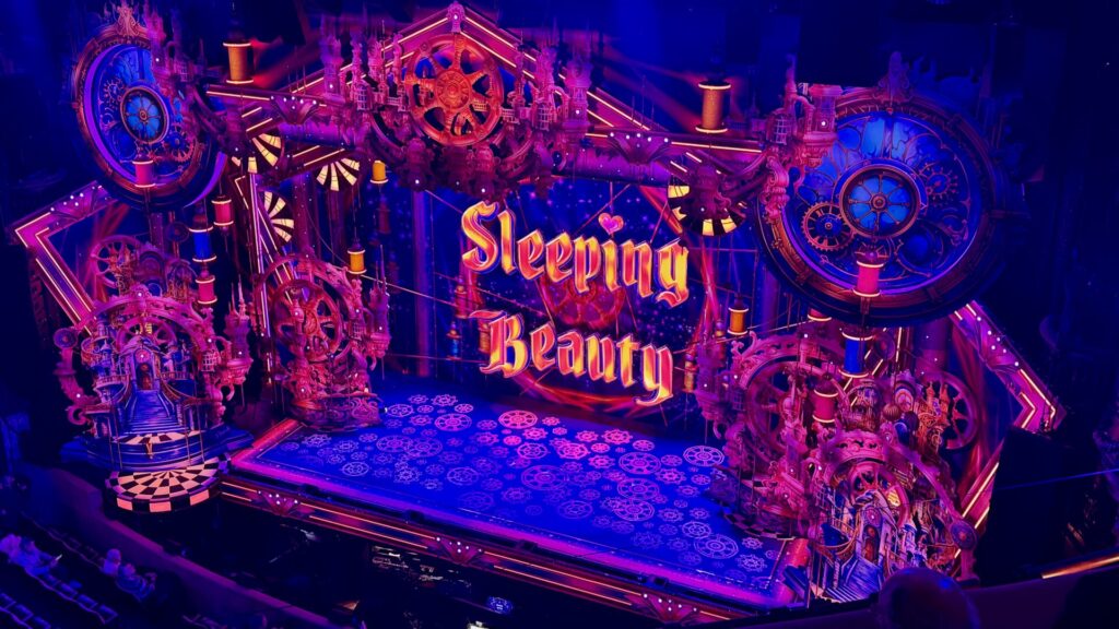 Elaborate theatrical stage set for 'Sleeping Beauty' pantomime illuminated in vibrant pink, purple, and blue lighting, featuring ornate spinning wheels, clock towers, thread spools, gears, and Gothic architectural elements