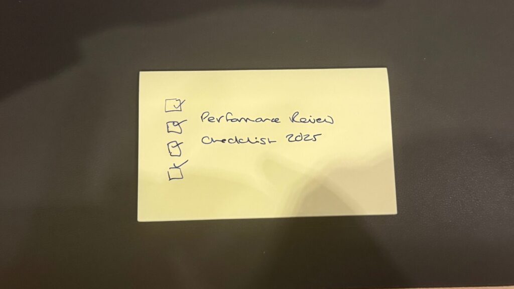 "A yellow sticky note on a dark grey surface with a handwritten checklist in blue ink. Four checkbox squares are drawn on the left side, with the first and fourth boxes ticked. The text reads 'Performance Review' and 'Checklist 2025'.