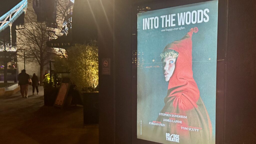 Illuminated poster for the Into the Woods musical at The Bridge Theatre, showing a stylised illustration of a figure in a bright coral-red hooded cloak against a teal-blue background with falling snowflakes. The figure has pale skin, blue eyes, and curled hair, gazing to the side. The poster displays the tagline 'and happy ever after' beneath the title, with credits showing music and lyrics by Stephen Sondheim, book by James Lapine, directed by Jordan Fein, and set and costume design by Tom Scutt. The Bridge Theatre logo appears at the bottom. The poster is mounted on a dark column in an evening street scene with trees and pedestrians visible in the background.
