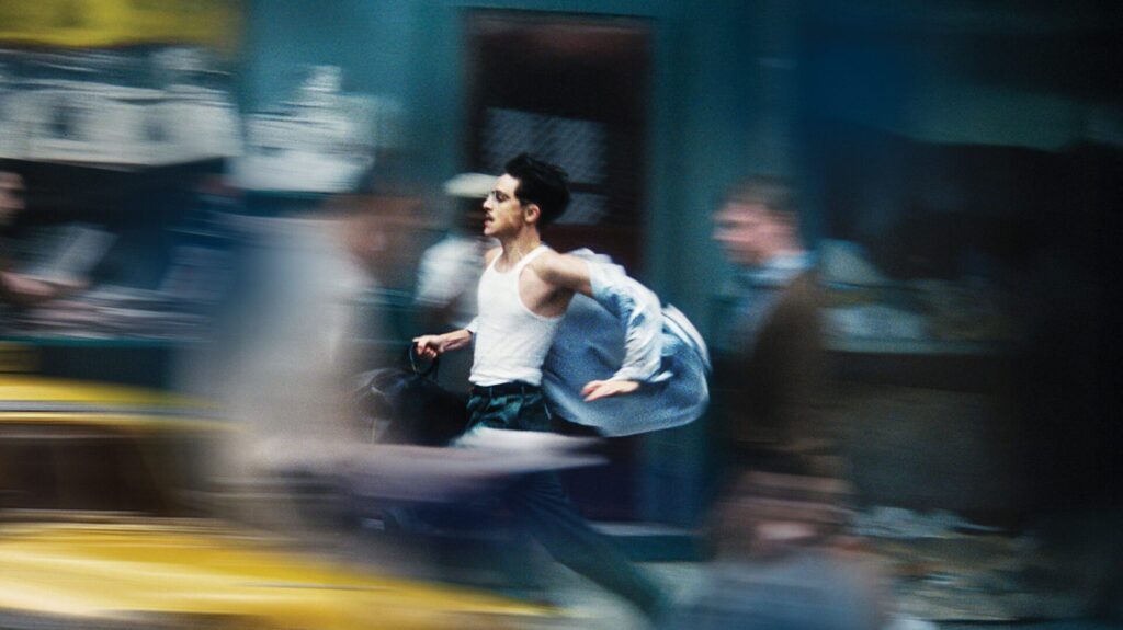 Motion-blurred photograph of a young man in a white vest and dark trousers mid-leap on an urban street, carrying a light blue jacket, with blurred vehicles and buildings in the background creating a sense of rapid movement © A24