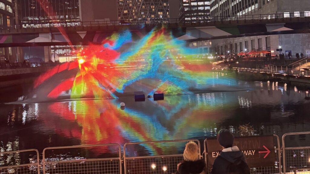 Colourful light installation reflected in water at Canary Wharf's Eden Dock at night. Vibrant rainbow beams of red, blue, yellow, and green light form geometric patterns across the water surface, with modern office buildings in the background and spectators viewing from behind barriers in the foreground.
