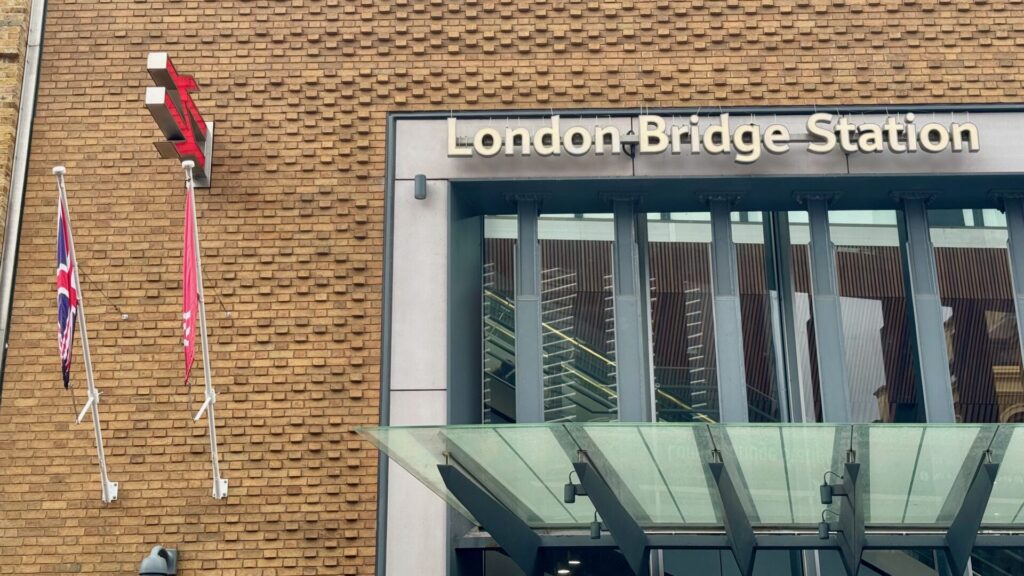 Modern entrance to London Bridge Station with glass canopy and metallic lettering on beige brick facade, Union Jack and British Rail flags visible on left
