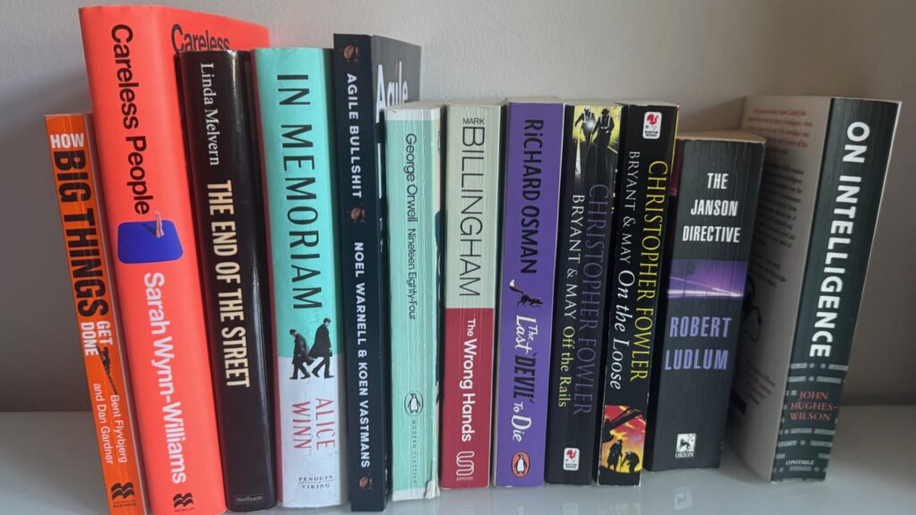 Row of thriller and spy novels on a bookshelf including titles by Sarah Vaughan-Williams, Linda Meiklem, Alice Winn, Noel Warrell, George Orwell, Mark Billingham, Richard Osman, Christopher Fowler, and Robert Ludlum, displaying varied spine colours from orange to black,
