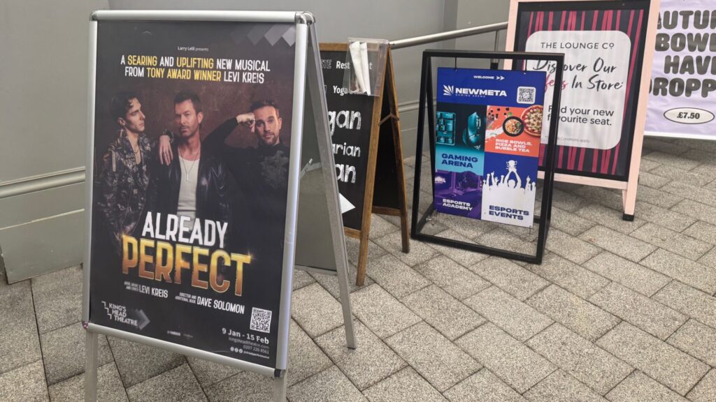 Promotional A-board sign for the musical 'Already Perfect' showing three male performers, positioned on a tiled floor alongside other advertising boards for a restaurant, gaming arena and bowling venue.