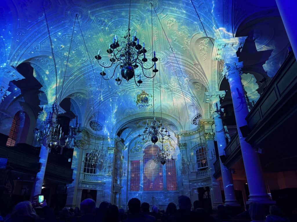 Interior of St Martin-in-the-Fields church filled with blue, green, and violet light projections during the Luxmuralis “Space” light and sound show, highlighting the ornate vaulted ceiling, chandeliers, classical columns, and a seated audience below.