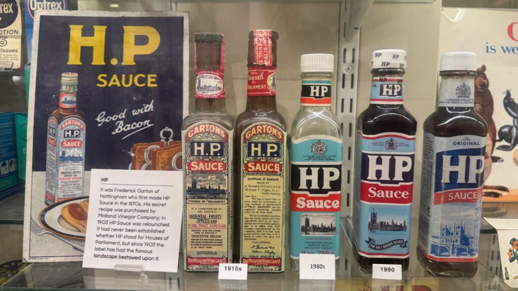 A museum display showing the evolution of HP Sauce packaging, featuring a vintage advertising card and five bottles spanning from the 1910s to 1990. The earliest bottles carry the "Garton's H.P. Sauce" label from the Midland Vinegar Company, with dense Victorian-era typography describing the sauce as a blend of oriental fruits, spices and pure malt vinegar. Later bottles show the progressive simplification of the label design, retaining the iconic Houses of Parliament illustration throughout. A printed card to the left notes that the sauce was first made by Frederick Garton of Nottingham in the 1870s, and that the recipe was sold to the Midland Vinegar Company in 1903. On display at the Museum of Brands, London.