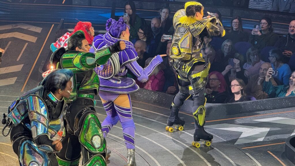 Performers in elaborate armoured costumes on roller skates during a live performance of Starlight Express at Wembley, London. Four characters are visible in close-up, each dressed in distinctive colour-coded outfits: one in iridescent blue, one in metallic green with matching shoulder armour, one in purple with striped detailing and a purple afro, and one in gold and yellow with a yellow mohawk. A crowd of audience members watches closely from behind a low barrier, several holding up phones to photograph the action. The stage floor features a curved track with geometric markings.