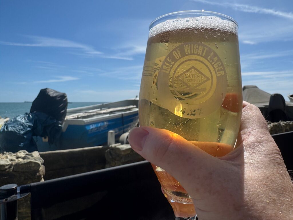 A hand holding a chilled glass of lager in bright sunshine outside The Fisherman’s Cottage pub in Shanklin, Isle of Wight, with fishing boats, the sea, and a clear blue sky in the background.