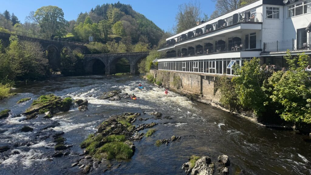 View from the Chainbridge, Llangollen, looking along the River Dee. Three kayakers navigate white-water rapids amongst rocky outcrops in the foreground. To the right, the white multi-storey Chainbridge Hotel sits alongside the riverbank, its tiered balconies and glass-fronted dining room overlooking the water. A stone arched bridge crosses the river in the middle distance, backed by a tree-covered hillside under a clear blue sky.