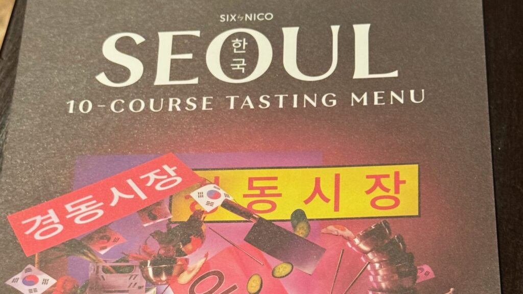 Menu cover for Six by Nico's Seoul-themed 10-course tasting menu, featuring bold white typography on a dark grey background with a vibrant collage illustration of Korean cultural imagery, including the South Korean flag, traditional armour, street food, and Korean signage reading "Gyeongdong Market".