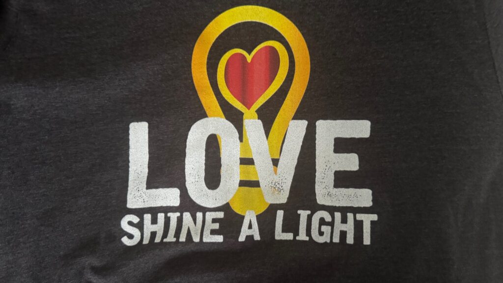 Close-up of a dark grey Eurovision Song Contest themed T-shirt. The design features a gold light bulb graphic in which the bulb's interior forms a red heart shape, with the filament stem extending below. In bold white text beneath the graphic are the words "LOVE" and "SHINE A LIGHT" — a reference to the UK's Eurovision-winning song performed by Katrina and the Waves in 1997.