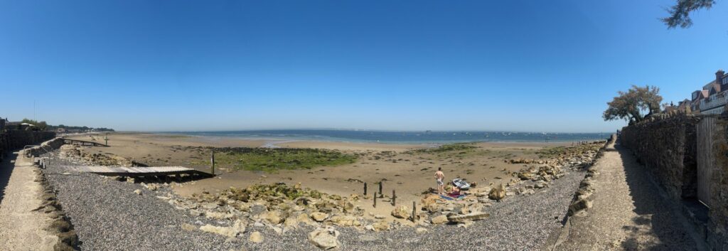 Panoramic view of a sandy beach at low tide on a sunny day in Seaview, Isle of Wight. Rocky gravel foreground leads to a wide expanse of sand dotted with seaweed and wooden posts, with calm blue water and boats visible in the distance. A lone figure stands beside a small dinghy. Residential properties and a sea wall frame the right side.