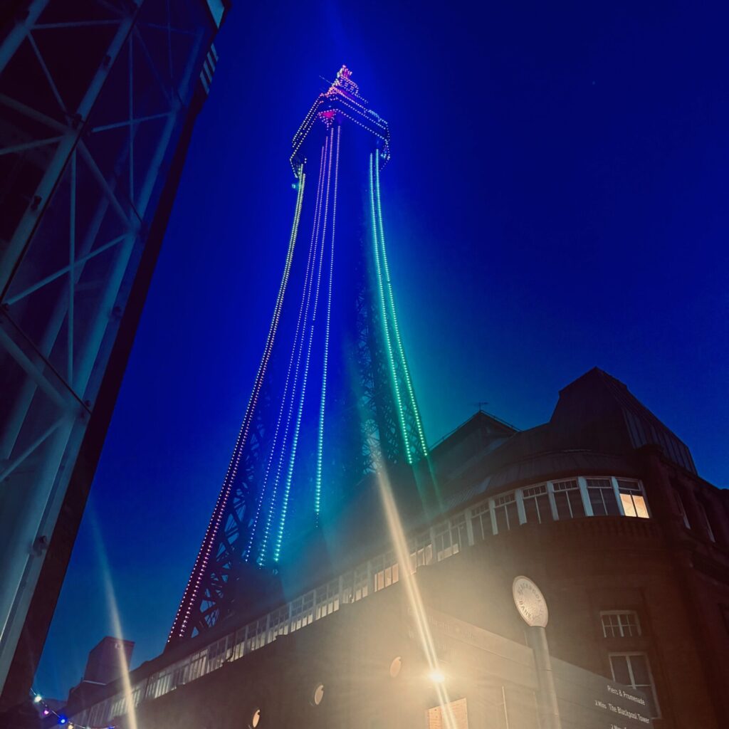 Blackpool Tower illuminated in rainbow Pride colours against a deep blue night sky, photographed from street level looking upward. Strings of LED lights in red, orange, yellow, green, blue and violet cascade down the tower's steel lattice structure. A warm streetlamp glows in the foreground, and the curved facade of a Victorian brick building is visible to the right, bearing a sign reading "Blackpool Promenade".