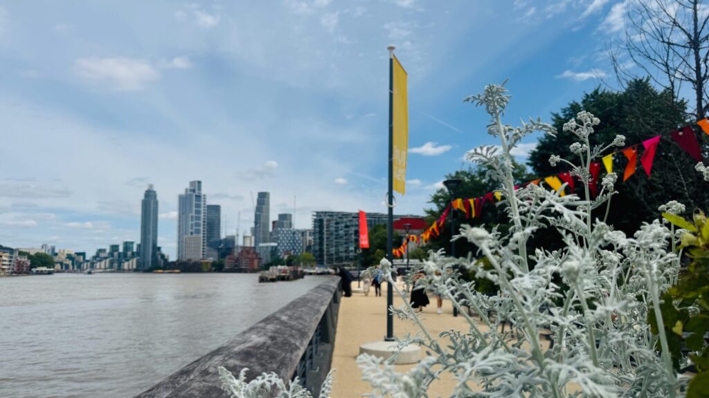 A riverside promenade along the Thames at Battersea Power Station, viewed on a partly cloudy summer's day. In the foreground, silvery-white dusty miller plants grow in raised planters beside a stone balustrade. A yellow banner on a lamppost and colourful bunting in red, orange and yellow hang along the walkway, where a few pedestrians stroll. Across the river, the Vauxhall skyline is visible, with modern high-rise residential towers rising against a blue sky scattered with white clouds.