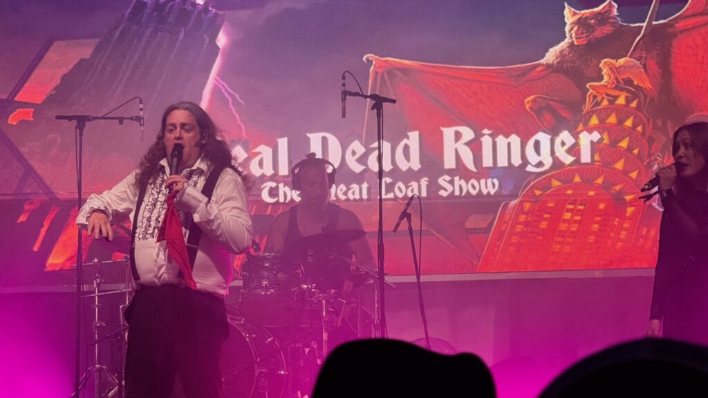 A long-haired male performer in a white ruffled shirt, black waistcoat and trousers holds a microphone and a red cloth on a purple-lit stage. Behind him, a drummer plays in the background, and a large screen displays the text "Real Dead Ringer – The Meat Loaf Show" alongside gothic artwork featuring a dramatic red sky and a snarling wolf.
