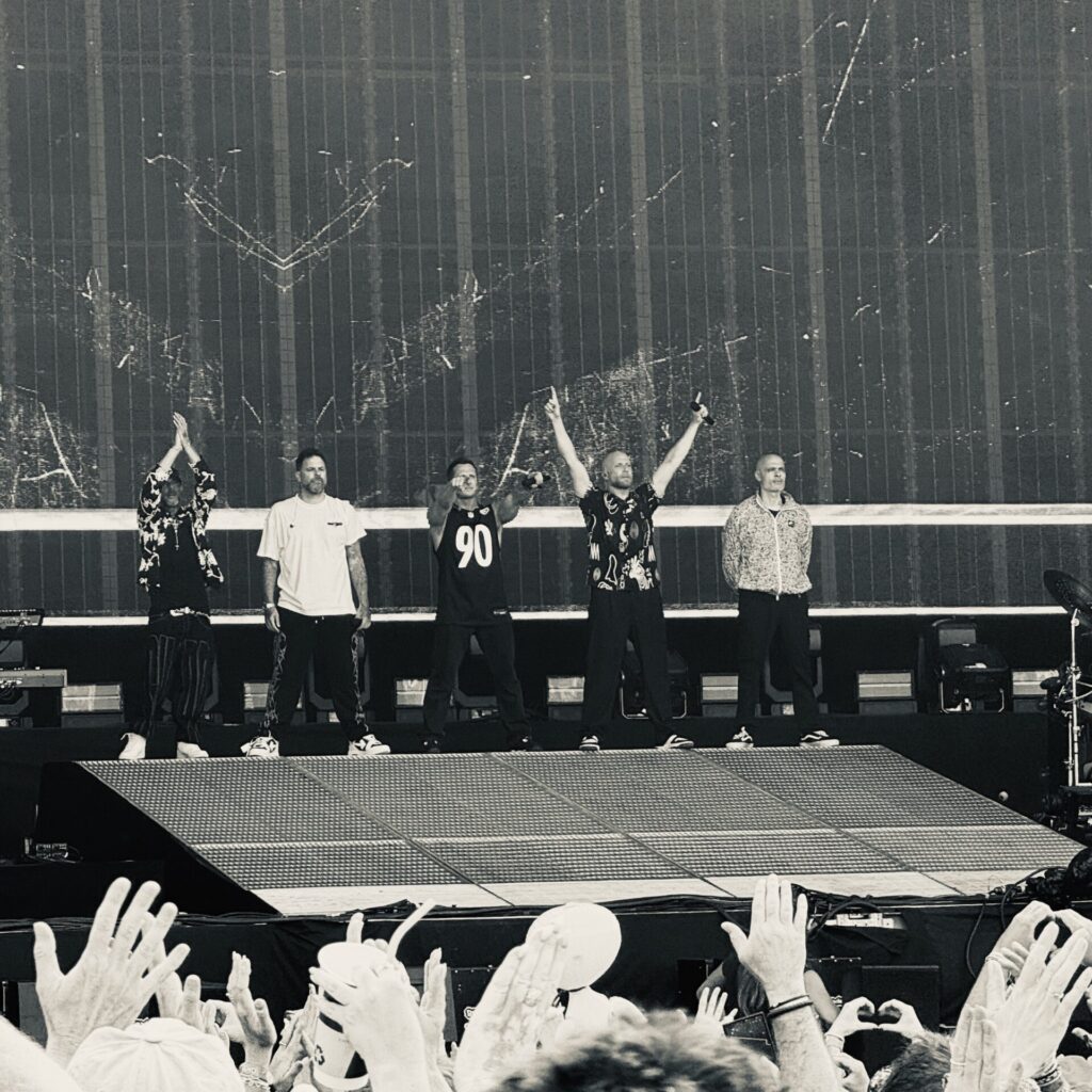 A black-and-white photograph of five men standing in a line on a large festival stage, acknowledging the crowd. Two members raise their arms, one wearing a black sports jersey with the number 90. The audience in the foreground has hands raised, some forming heart shapes. A large LED screen with an abstract scratched pattern forms the backdrop.