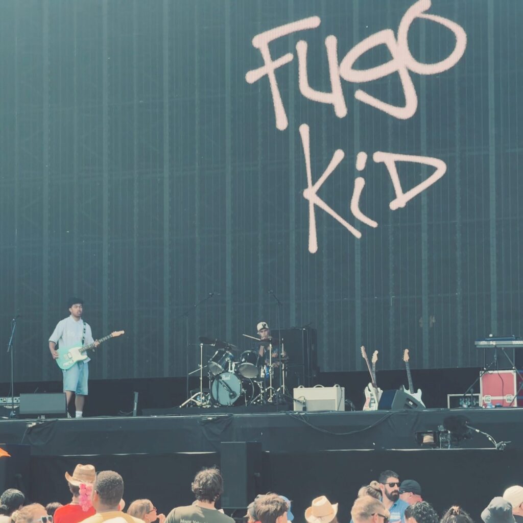 Fugo Kid performs on a large outdoor festival stage. A guitarist in a pale blue oversized shirt and denim shorts plays a light blue electric guitar on the left, whilst the drummer in a white cap sits behind a black kit at the centre rear. Two spare electric guitars on stands are visible to the right of the stage. The band's name is displayed in loose, hand-drawn pink lettering on the large dark backdrop screen. The crowd below is visible in the foreground.