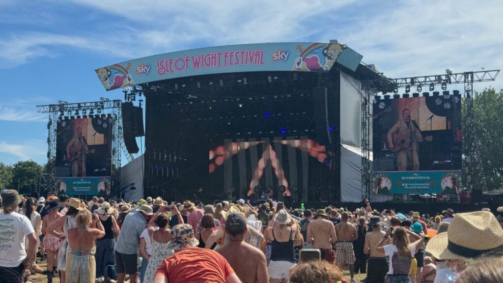 A large sun-drenched crowd faces the Sky-sponsored Isle of Wight Festival main stage during David Gray's afternoon performance. The central LED screen displays abstract warm-toned imagery, whilst the two flanking side screens show close-up footage of David Gray performing in a cream suit and holding an acoustic guitar. Festivalgoers in summer clothing, hats, and bare shoulders fill the foreground beneath a bright blue sky dotted with clouds.