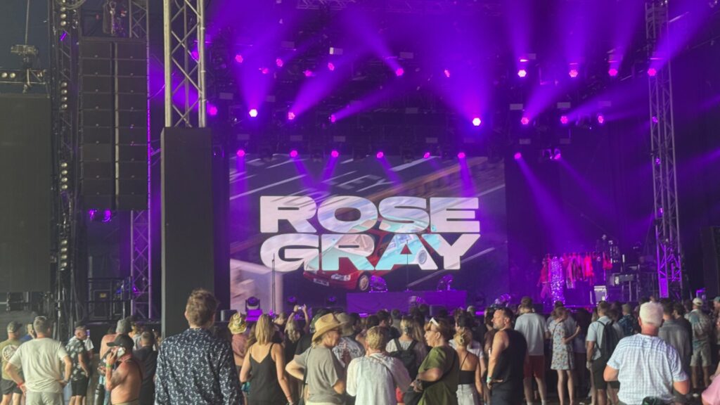 A festival stage bathed in deep purple lighting, with a large LED screen displaying "Rose Gray" in bold white block lettering over a backdrop of a blurred fast-moving road. Stage lights beam outward in vivid purple rays above the screen. A gathering crowd stands in the foreground facing the stage, with guitars on stands visible to the right awaiting the performance.