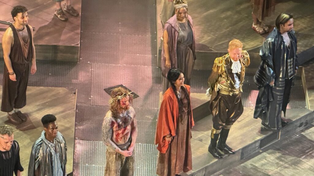 Cast members of Jesus Christ Superstar take their bows on stage at the London Palladium. Photographed from above, the ensemble includes Sam Ryer in the role of Jesus wearing a crown of thorns and a blood-streaked costume, Jesse Tyler Ferguson in an ornate gold military-style jacket representing Herod, a woman in an orange robe, and several other cast members in period and contemporary costumes, all lined up across the stage.