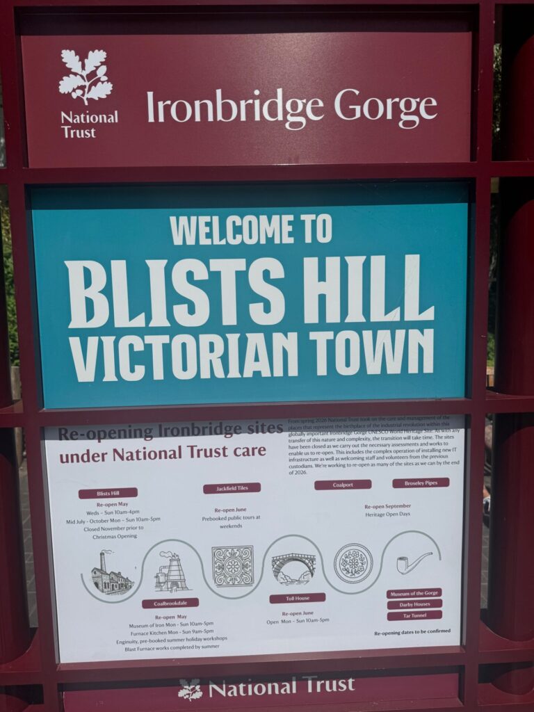 The entrance sign for Blists Hill Victorian Town, part of the Ironbridge Gorge UNESCO World Heritage Site, now under National Trust stewardship. The sign features the National Trust oak leaf logo at top left alongside the text "Ironbridge Gorge" on a dark red background. Below, a large teal panel reads "Welcome to Blists Hill Victorian Town" in bold white lettering. A lower white panel headed "Re-opening Ironbridge sites under National Trust care" lists reopening schedules for several sites including Blists Hill, Coalbrookdale, Jackfield Tiles, Coalport, Toll House, Broseley Pipes, and others, with dates ranging from May to September 2026. Small illustrated icons represent each attraction. The National Trust logo appears again at the foot of the sign.