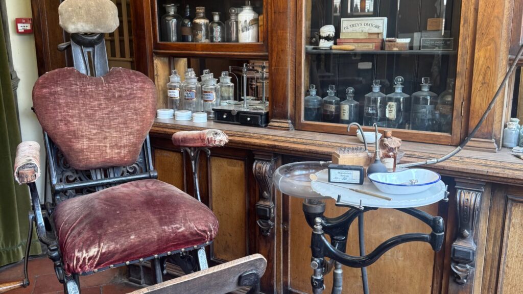 A Victorian dentist's chair on display at Blists Hill Victorian Town museum, now under National Trust stewardship. The ornate black cast-iron chair is upholstered in worn burgundy velvet with padded armrests and a shaped headrest. Beside it stands a small circular dentist's tray on a decorative iron stand, holding instruments, including a labelled "Hypodermic Syringes" and a white enamel spittoon bowl. Behind the chair, a dark wooden cabinet and worktop are lined with rows of glass apothecary bottles, small white ceramic pots, and period dental products, including a box labelled "De Trey's Diatoric" and a tin of toothpaste.