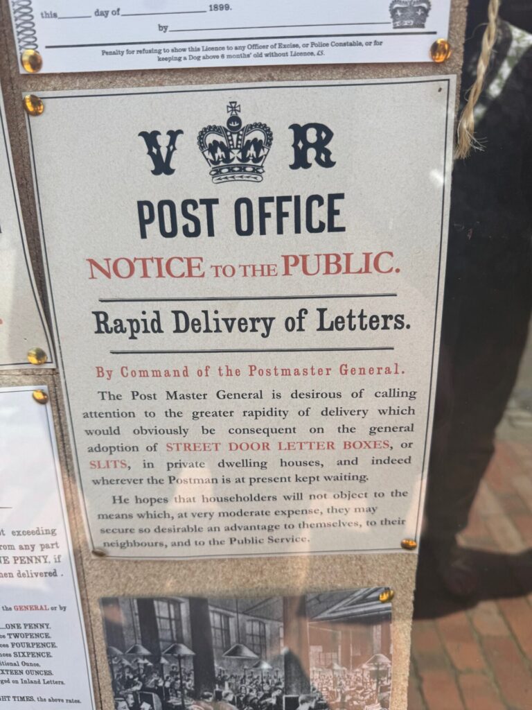 A reproduction Victorian Post Office public notice, displayed on a noticeboard at Blists Hill Victorian Town museum. Headed with the royal cypher "V R" and a crown, the notice is titled "Post Office — Notice to the Public: Rapid Delivery of Letters — By Command of the Postmaster General." The text urges householders to install street-door letter boxes, or "slits," in private dwellings to speed up postal delivery and avoid keeping the postman waiting. The notice is printed in a mix of black and red Victorian typefaces on cream paper and fixed to a cork board with brass-effect studs. A partial view of other period notices is visible at the edges.