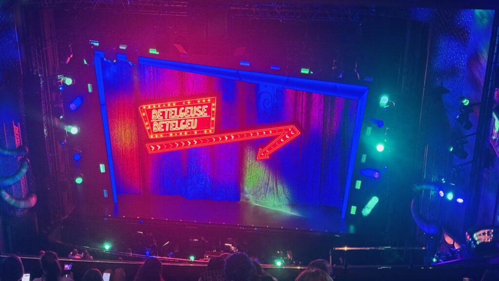 A theatre stage set photographed during a performance of Beetlejuice The Musical, lit in vivid pink, blue, and green stage lighting. At the centre of the stage hangs an illuminated red neon sign reading "Betelgeuse," styled like a vintage marquee, with a flashing chevron arrow beneath it pointing diagonally downward. A large blue rectangular frame structure surrounds the scene, with patterned dark curtains visible behind. Silhouettes of audience members' heads are visible in the foreground, with some holding up phones to photograph the stage.