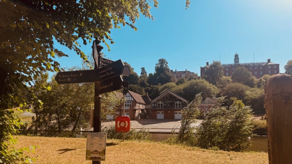 A Victorian cast-iron fingerpost in Shrewsbury's Quarry Park, signposted for St Julian Friars, the Swimming Centre, Children's Play Area and Town Centre, with the riverside boathouse and pavilion below and Shrewsbury's rooftops and a clock tower visible beyond the trees on a clear summer day.