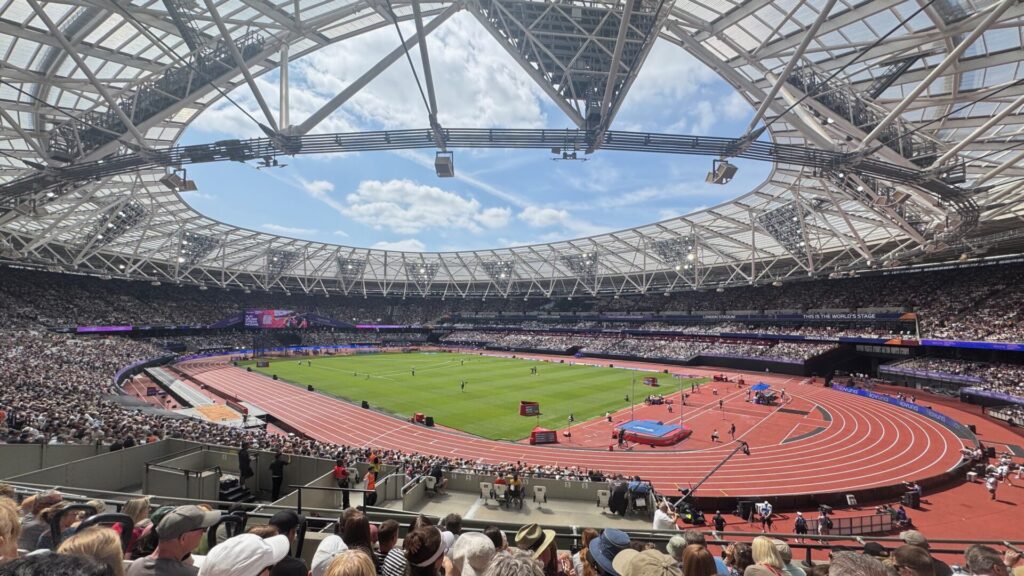 A wide-angle view inside London Stadium during the Novuna London Athletics Meet, showing the packed stands, the running track and infield with athletes preparing for a field event, all beneath the stadium's distinctive open roof structure against a blue sky.
