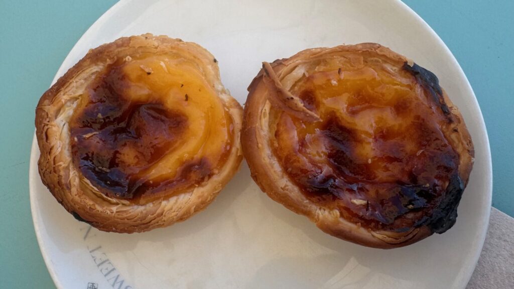 Two golden pastéis de nata on a white plate, their flaky pastry cases filled with custard that's caramelised dark brown in places, one showing a slightly more scorched top than the other.