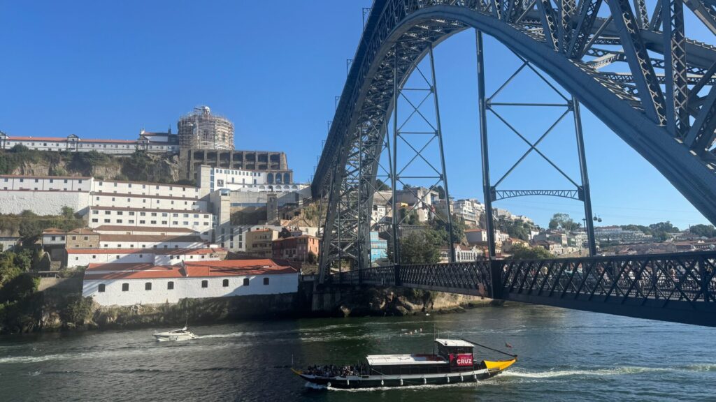 A view of the Dom Luís I Bridge in Porto, its iron double-deck arch framing the whitewashed port wine cellars of Vila Nova de Gaia and the hillside monastery above, with a traditional rabelo boat and a motor yacht passing on the Douro River below.