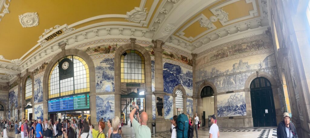 The ornate main hall of São Bento Railway Station in Porto, its walls covered in blue-and-white azulejo tiles depicting historical scenes, beneath a gilded, sculpted ceiling, with an arrivals board and travellers gathered below.