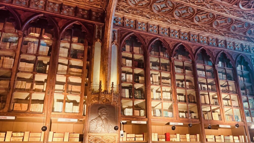 Ornate carved wooden shelving inside Livraria Lello in Porto, filled with Spanish-language books behind glass-fronted Gothic arches, with a bronze relief plaque set into a central column and an intricately carved ceiling above.
