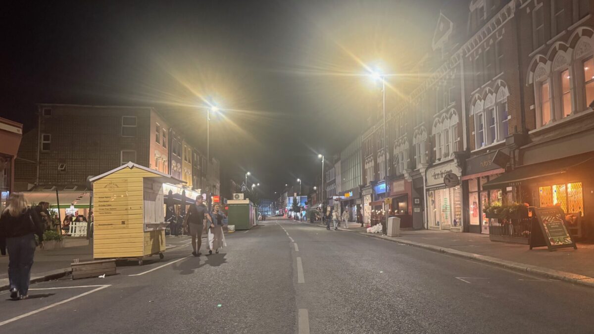 Northcote Road in Battersea photographed at night, closed to traffic for the weekend and largely empty, with streetlights glaring overhead, shopfronts lit along both sides, and a couple walking past a wooden market stall.