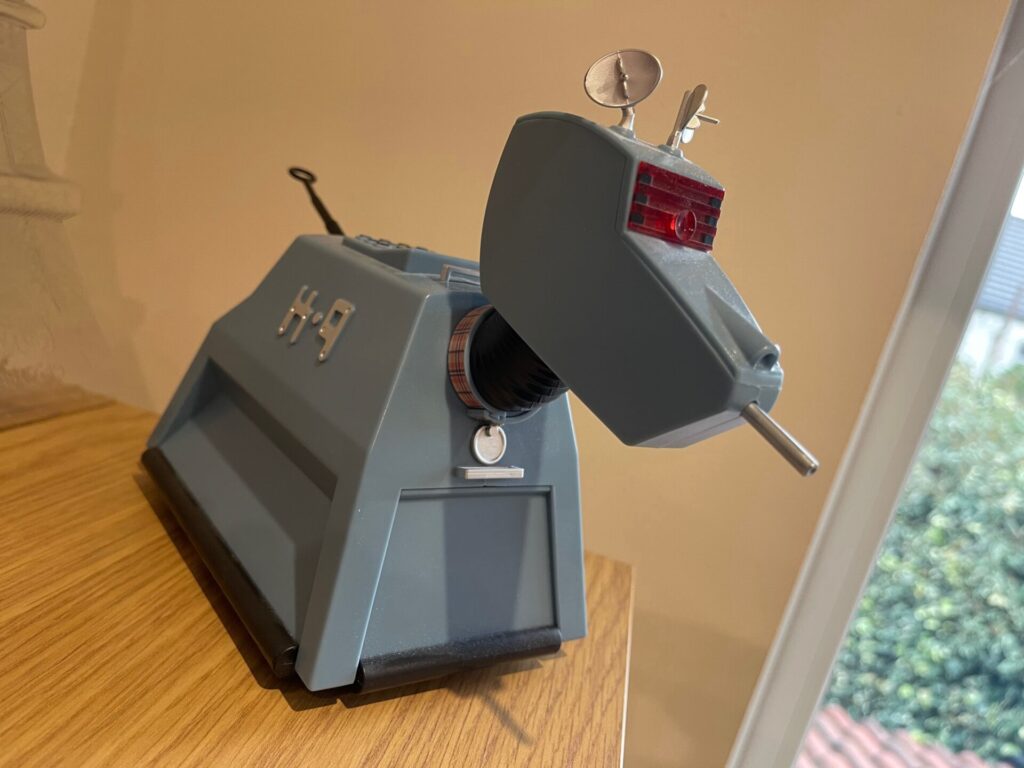 A detailed K9 model from Doctor Who, displayed on a wooden table, its head tilted upward on an extended neck, with its trademark ear-like radar dishes, red laser-tipped nose gun and tail-mounted probe visible against a warm-lit room.