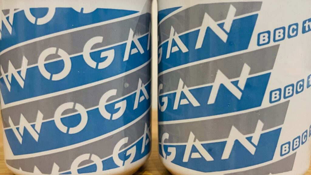 Two white mugs printed with the repeated word "WOGAN" in diagonal blue and grey bands alongside a small "BBC1" logo, photographed in close-up.