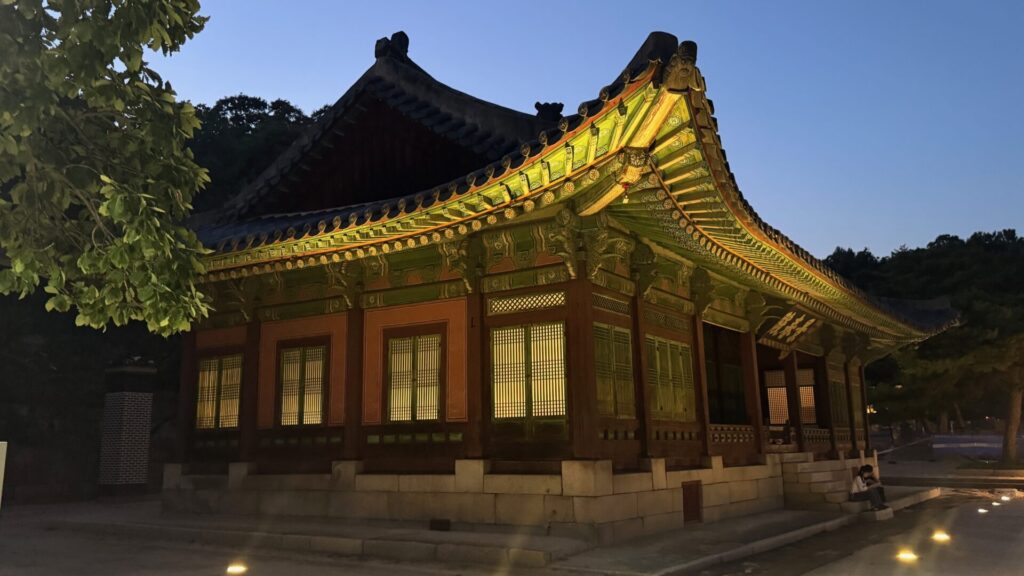 A traditional Joseon-era hall at Changgyeonggung Palace in Seoul at dusk, its sweeping tiled roof and richly painted dancheong eaves lit in glowing green and gold, with warm light shining through the lattice windows and a visitor sitting on the stone steps on the right.
