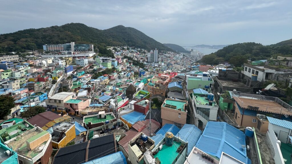 Panoramic view of Gamcheon Culture Village in Busan, South Korea, showing densely packed, colourful houses with blue-and-green rooftops cascading down a hillside towards the harbour, surrounded by wooded mountains.