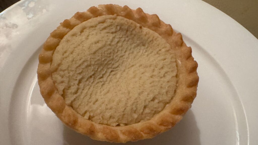 Close-up of a mince pie on a white plate, with a golden-brown crimped pastry edge surrounding a pale, textured pastry top.